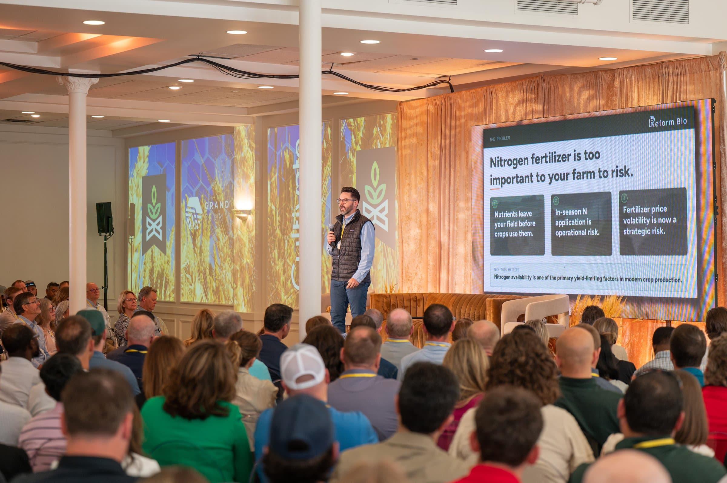 Kyle Anderson speaking on stage at a Grand Farm 'Cultivate' event, with Grand Farm branding and a Reform Bio presentation slide visible, in front of a large seated audience.